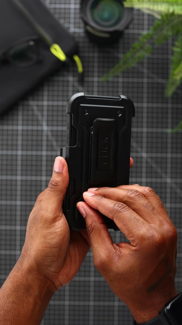 An iPhone 17 Pro in a black CLICK Latch Holster case, seated in its belt-clip holster and held in two hands